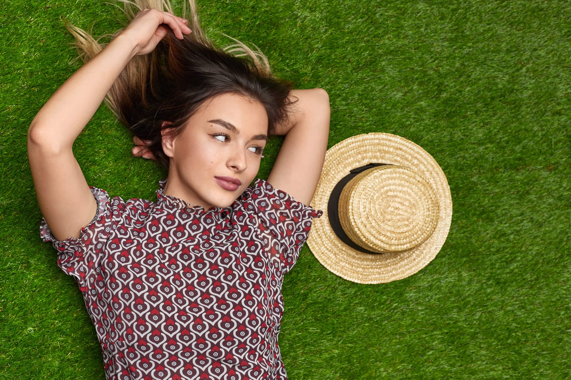 A young woman with dark hair and blonde highlights lies on green grass. She's wearing a red and white patterned top and has her arms raised above her head in a relaxed pose. A straw sun hat is placed next to her on the grass. Her expression is serene as she looks to the side, creating a peaceful, summery atmosphere.