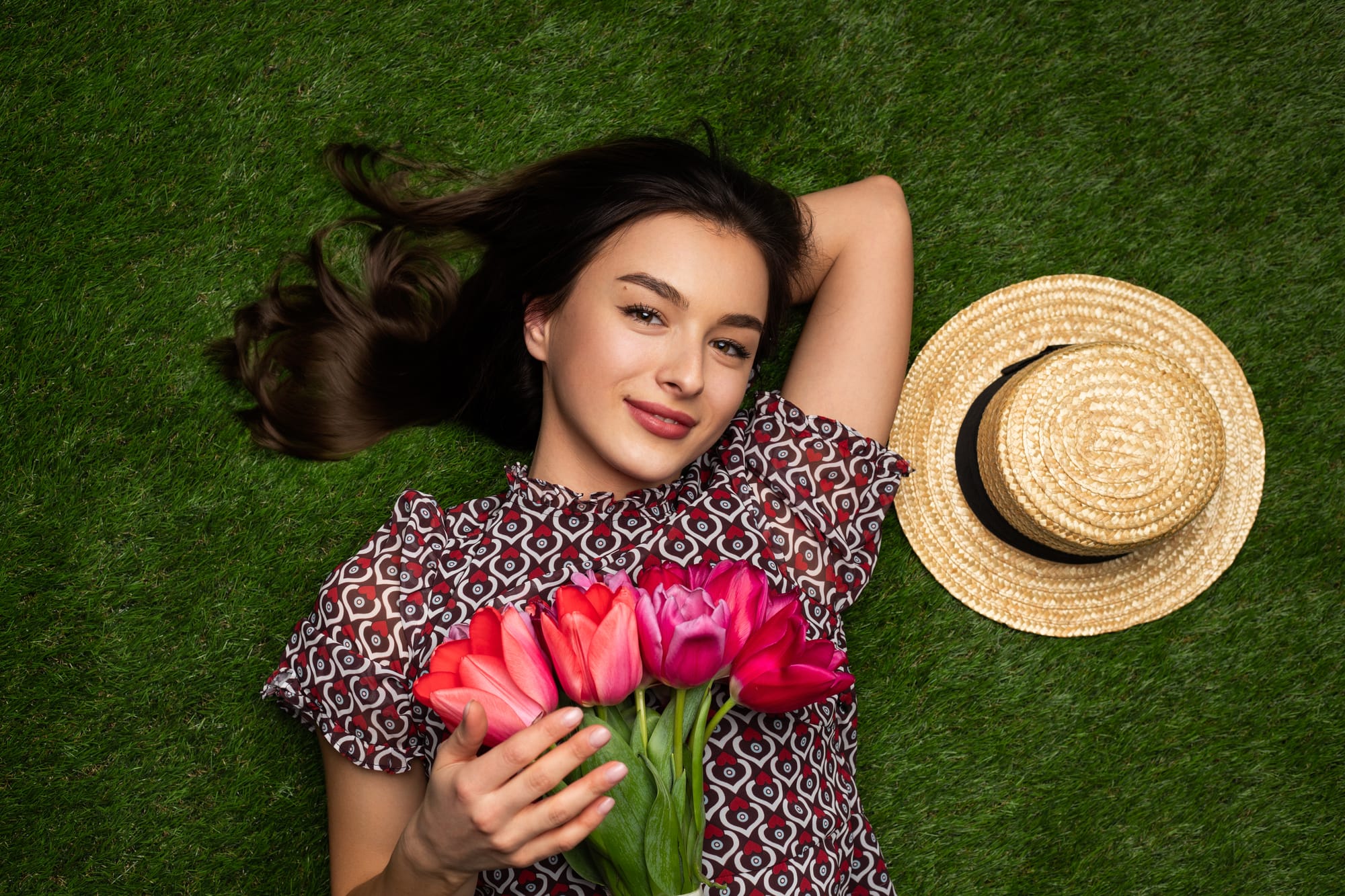 A young woman with long dark hair lies on green grass, smiling at the camera. She's wearing a red and white patterned top and holding a bouquet of pink and red tulips against her chest. A straw sun hat is placed next to her head. The image conveys a cheerful, springtime atmosphere.