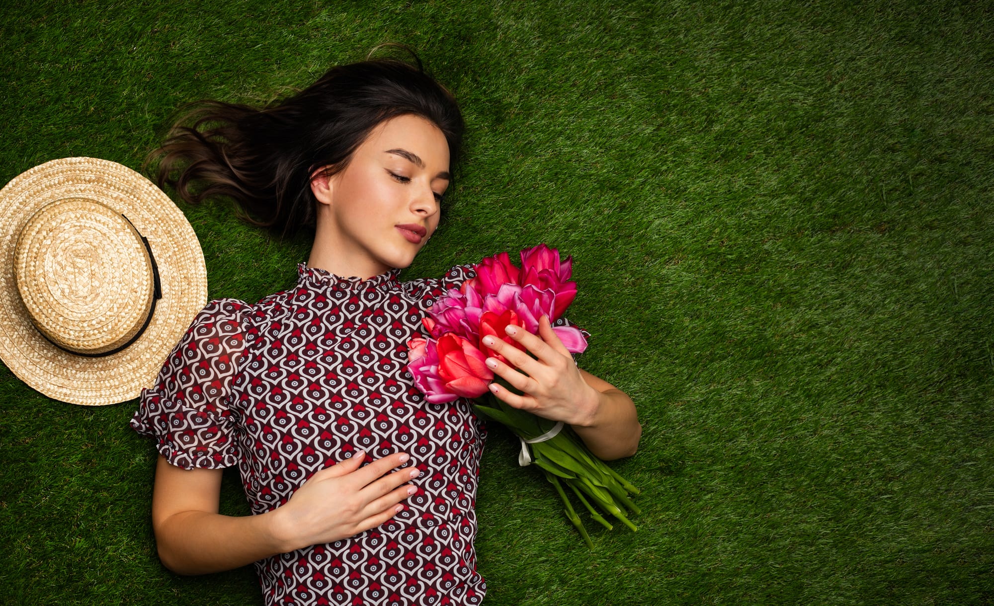 A young woman with dark hair lies on green grass with her eyes closed, holding a bouquet of vibrant pink tulips to her chest. She's wearing a red and white patterned top with short sleeves. A straw sun hat is placed next to her head. The image evokes a peaceful, springtime mood.